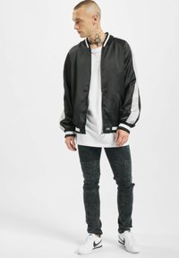 Lett svart bomberjakke med hvite striper, kombinert med en lang hvit skjorte, slitte svarte jeans og hvite sneakers.