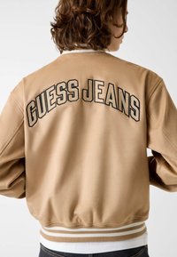 Beige Bomberjacke mit "GUESS JEANS" in Schwarz gestickt, weiß geripptem Besatz am Kragen und Saum, mit glattem Satin-Finish.