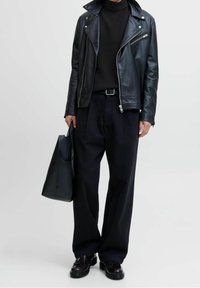 Veste de motard en cuir noir avec zips, col roulé côtelé noir, pantalon noir ample et chaussures noires brillantes. Sac cabas noir à la main.