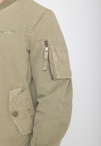 Khaki jas mouw met ritssluiting en klepzak, geborduurd gevleugeld embleem nabij de schouder en een onderste zak met knoop.