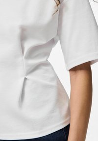 Bijela majica kratkih rukava od mekog materijala, s bočnim nabiranjima i zaobljenim rubom. Vidljive detalje šavova. Nosiva s tamnim trapericama.