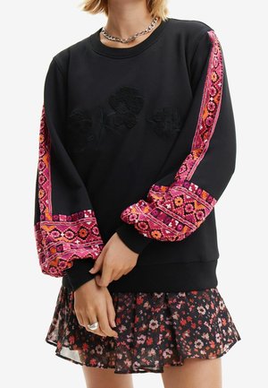 Femme portant un sweat-shirt noir avec des appliqués floraux noirs et des manches brodées rose vif, associé à une jupe fleurie noire et un collier à chaîne argentée.