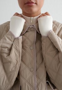 Beige quiltet jakke med diamantmønster, med høy krage og kontrastdetaljer. Inkluderer hvite, tettsittende mansjetter for varme.