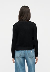 Γυναίκα με μακριά σκούρα μαλλιά που φοράει μαύρο μακρυμάνικο τοπ με υφή και ανοιχτό μπλε τζιν παντελόνι, όρθια με την πλάτη προς την κάμερα.