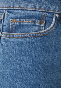 Tela de mezclilla azul de unos vaqueros con un bolsillo trasero con costuras naranjas y una pequeña etiqueta blanca. Superficie texturizada con áreas descoloridas.