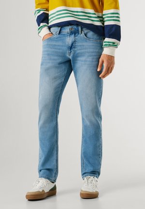 Mann trägt hellblaue Jeans, weiße Sneakers mit braunen Sohlen und ein gestreiftes Langarmshirt in Gelb, Blau und Grün.