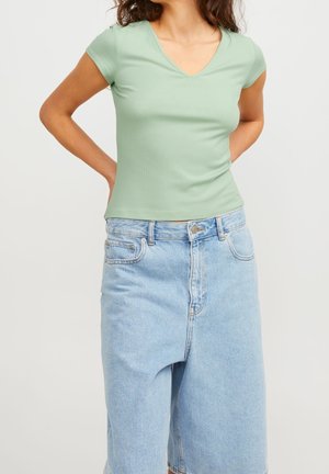 Vrouw draagt een lichtgroene korte mouw V-hals top en hooggetailleerde losse lichtblauwe denimshorts, staand met handen achter de rug.