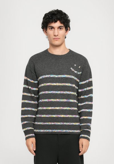 Uomo con capelli ricci che indossa un maglione grigio scuro con strisce orizzontali multicolori e un piccolo motivo di faccina sorridente sul petto.