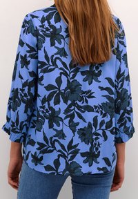 Femme aux longs cheveux portant un chemisier bleu à motifs floraux foncés et un jean bleu clair, debout, dos à la caméra.