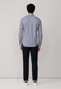 Hombre de pie frente a una pared, vistiendo una camisa de manga larga a cuadros azul, jeans oscuros arremangados y zapatillas blancas.
