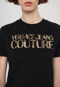 Črna bombažna majica s kratkimi rokavi z bleščečim zlatim napisem "VERSACE JEANS COUTURE" čez prsi. Oblika s krojnim ovratnikom.