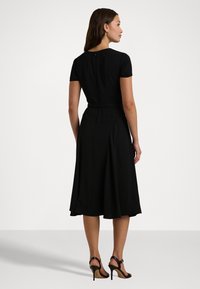 Μαύρο φόρεμα με περιτύλιγμα, κοντά μανίκια, εφαρμοστή μέση, φαρδύ φούστα και φερμουάρ στην πλάτη. Φορεμένο με ψηλοτάκουνα παπούτσια με λουριά.