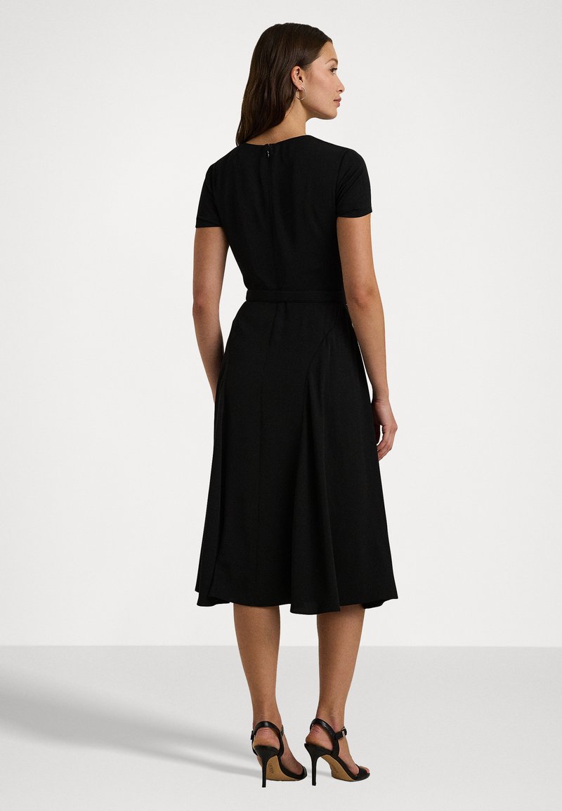 Abito nero avvolgente con maniche corte, corpetto aderente, gonna a campana e zip sul retro. Indossato con scarpe con tacco a strappo.