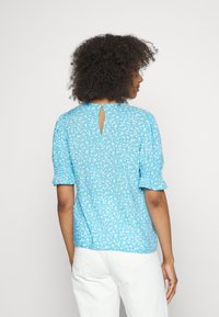 Blusa floral azul com mangas bufantes e detalhe em forma de buraco na parte de trás. Tecido leve com uma textura suave e pequeno padrão floral branco.