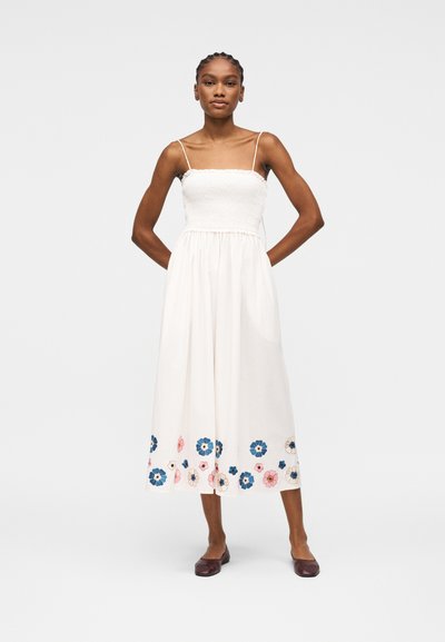 Femme portant une robe midi blanche sans manches avec un corsage froncé et une broderie florale colorée le long de l'ourlet, assortie à des ballerines foncées.