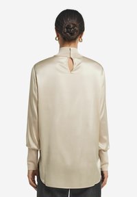 Satinbeige blus med hög krage, nyckelhålsdetalj i ryggen, långa ärmar och sidoklyftor vid fållen för ökad rörlighet.