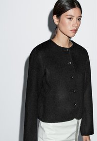 Kvinna med mörkt hår uppsatt i en svart knäppt jacka och vit kjol, vänd mot höger mot en ljus, enkel bakgrund.