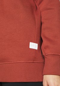 Röd sweatshirt med ribbade muddar, flatlock-sömmar och en fyrkantig vit etikett som har "G-STAR RAW" på nedre vänstra sidan.