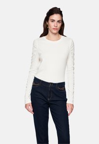 White long-sleeve top with gathered detailing along the sleeves, paired with dark blue jeans featuring subtle stitching and pockets.