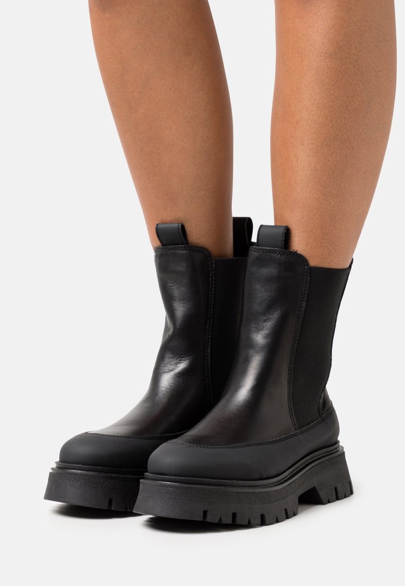 Bottines en cuir noir avec semelles épaisses à crampons portées sur des jambes nues sur un fond blanc uni.