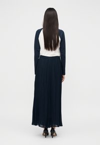 Μπλε σκούρα πλισέ φούστα με ελαστική ζώνη στη μέση, συνδυασμένη με λευκό και μπλε μακρυμάνικο τοπ. Το μοντέλο φοράει μαύρα παπούτσια με ψηλό τακούνι.