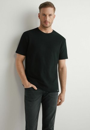 Mężczyzna z krótkimi włosami ubrany w czarny, teksturowany t-shirt z krótkim rękawem i ciemne jeansy stoi oparty o białą ścianę, z jedną ręką w kieszeni.