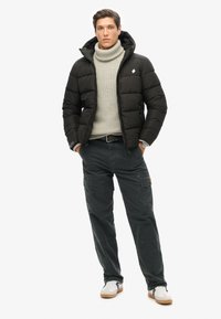 Sort pufferjakke med hætte, grå turtleneck, mørke cargo-bukser og hvide sneakers. Jakken har logo. Glatte teksturer.