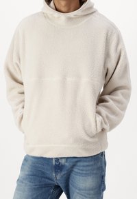 Pullover in pile di colore avorio, con colletto alto e grande tasca a marsupio. La texture è morbida e soffice con un fit comodo.