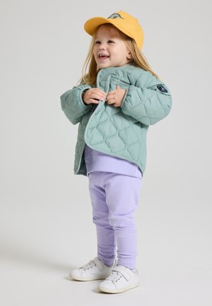 Gevoerde mintgroene gewatteerde jas, lila leggings en witte sneakers. Gele pet met een grafisch ontwerp voegt kleuraccenten toe aan de outfit.