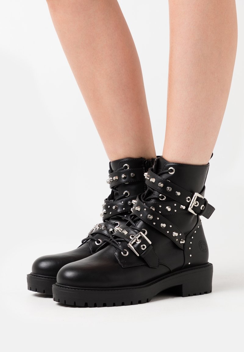 Bottes noires en cuir à la cheville avec plusieurs sangles cloutées et boucles argentées, portées sur des jambes nues sur fond blanc.