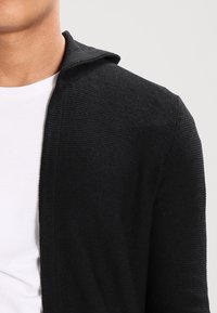 Černý pletený cardigan s kapucí, s texturovanou tkaninou a otevřeným předním designem, nošený přes bílé tričko.