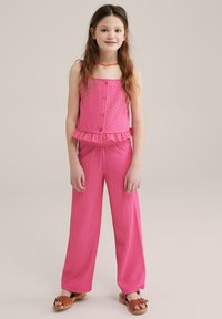 Rozā divdaļīgs apģērbs, kas sastāv no bezpiedurkņu topa ar pogām un volāna maliņu, kombinēts ar platām biksēm. Tekstūrēta auduma un ikdienišķas sandales.