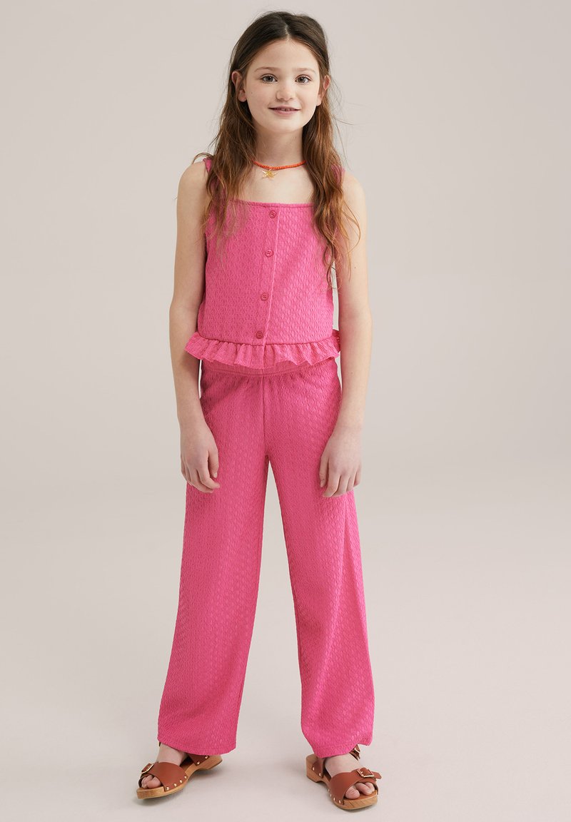 Rozā divdaļīgs apģērbs, kas sastāv no bezpiedurkņu topa ar pogām un volāna maliņu, kombinēts ar platām biksēm. Tekstūrēta auduma un ikdienišķas sandales.