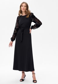 Schwarzes Maxikleid mit langen transparenten gemusterten Ärmeln, körperbetontem Oberteil, gebundenem Taillengürtel und glatter Stofftextur. Hochhackige Sandalen.