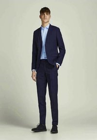 Jovem em pé, vestindo um fato azul escuro, camisa de vestir azul claro e sapatos de vestir pretos, diante de um fundo cinza liso.
