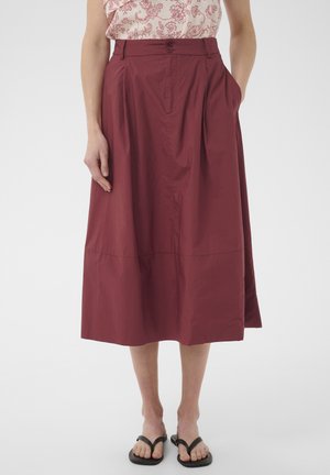 Femme portant une jupe midi bordeaux taille haute à boutons avec poches latérales, associée à des sandales noires en tongs et un haut à motifs.