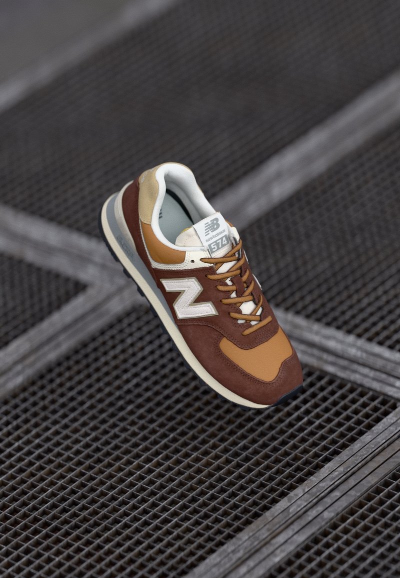 Baskets New Balance 574 marron et beige avec des accents blancs et des lacets blancs, positionnées au-dessus d'une surface métallique grillagée.