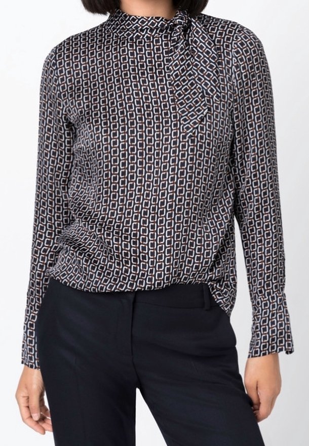 Femme portant un chemisier à manches longues avec un motif chaîne géométrique noir et blanc, rentrée dans un pantalon noir, debout devant un fond uni.