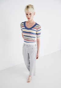 Večbarvna pletena majica s horizontalnimi črtami v modri, rdeči, zeleni in rumeni barvi, v kombinaciji s svetlo sivo širokimi hlačami in natikači.