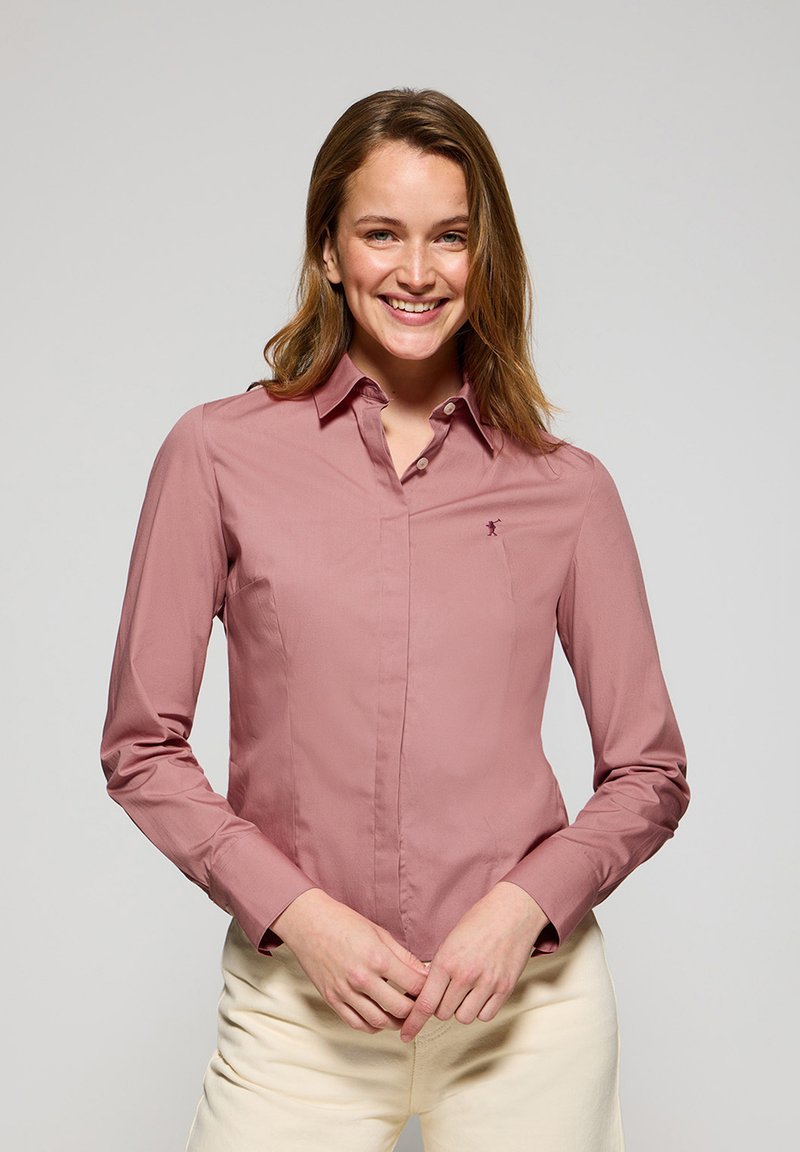 Camicia a maniche lunghe con colletto di colore rosa chiaro, dotata di una patta frontale e un piccolo logo sul lato sinistro del petto. Tessuto morbido e leggero.