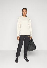 Krem sweatshirt, svarte joggebukser med gulllogo, svart veske og svarte sneakers. Enkelt design, avslappet passform og myk tekstur.