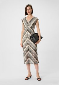 V-förmiges Kleid in Creme, Schwarz und Beige mit kurzen Ärmeln, knielangem Saum und Chevron-Design; kombiniert mit einer schwarzen Clutch und Mules.