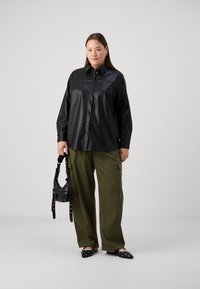 Črna bluza iz umetnega usnja z gumbnim zapiranjem, širokimi rokavi in dekorativnim šivom. Ujemajoča se olivno zelena cargo hlače in črna torbica.