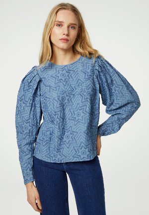 Blaue gemusterte Bluse mit Sternen, langen Puffärmel und rundem Ausschnitt. Eine Hose aus Denim ist sichtbar.