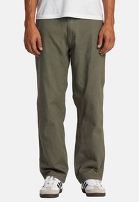Olive green cotton pants with a relaxed fit, straight legs, front pockets, and visible stitching; worn with white and black athletic shoes.