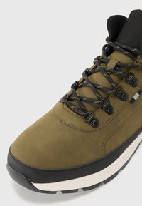 Olive green sneaker with a textured, synthetic upper, black accents, chunky white midsole, and sturdy black rubber outsole with tread pattern.