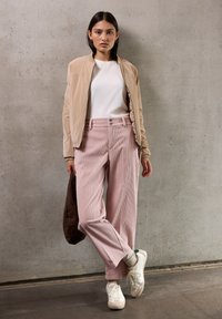 Donna in piedi contro un muro di cemento, indossa una giacca bomber beige, una camicia bianca, pantaloni ampi rosa, sneaker bianche e tiene in mano una borsa marrone.