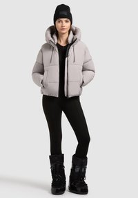 Giubbotto imbottito grigio chiaro con colletto alto e chiusura con zip. Leggings neri e stivali da neve neri alti completano l'outfit.