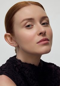 Złote minimalistyczne kolczyki w kształcie gałązki, noszone przez modelkę o jasnej karnacji i gładkim, naturalnym makijażu na neutralnym tle.