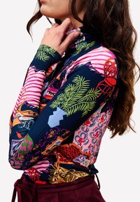 Frau mit langen lockigen Haaren, die einen bunten, gemusterten Rollkragenpullover mit floralen, gestreiften und abstrakten Designs trägt und den Kragen nahe ihres Halses hält.